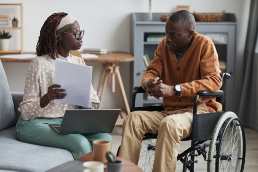 Portrait of a woman with a computer on her lap sharing a paper with an older African-American man utilizing a wheelchair