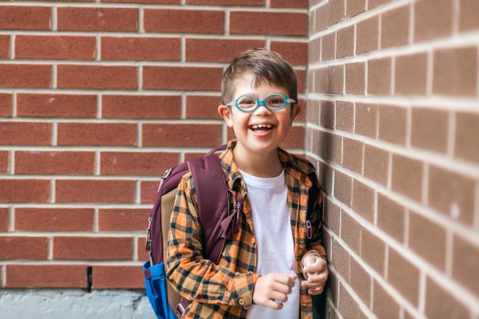 A child boy wearing glasses and a backpack poses next to a brick wall of the school