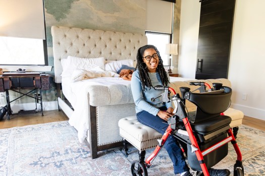 A portrait of a cheerful young adult woman with cerebral palsy sitting on the bench at the foot of her bed.