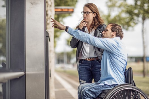 A man utilizing a wheelchair pointing at something on a building, explaining to a woman